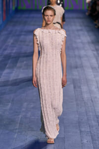 Model walking the runway in a pale pink, off-shoulder maxi dress with ruched, vertical detailing and ruffled neckline.