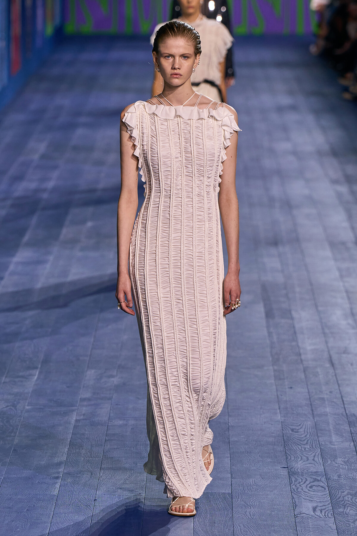 Model walking the runway in a pale pink, off-shoulder maxi dress with ruched, vertical detailing and ruffled neckline.