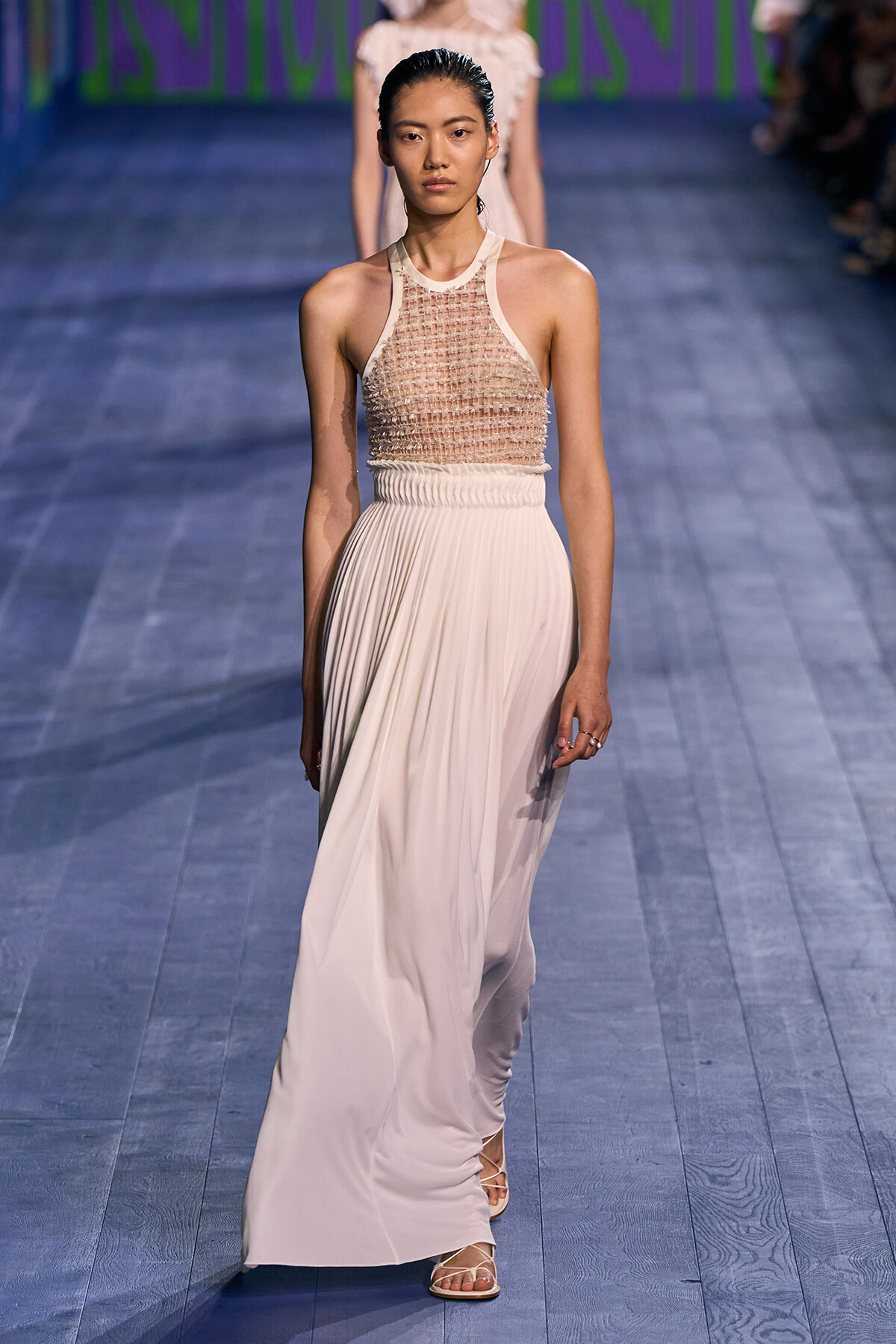 Model walking the runway in a beige, pleated, floor-length skirt with a beaded mesh halter top.
