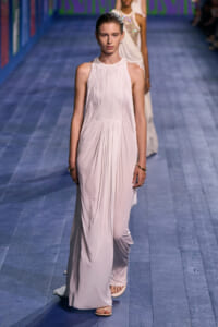 Model walking a pale pink, pleated halter-neck gown down a blue runway; minimal jewelry visible.