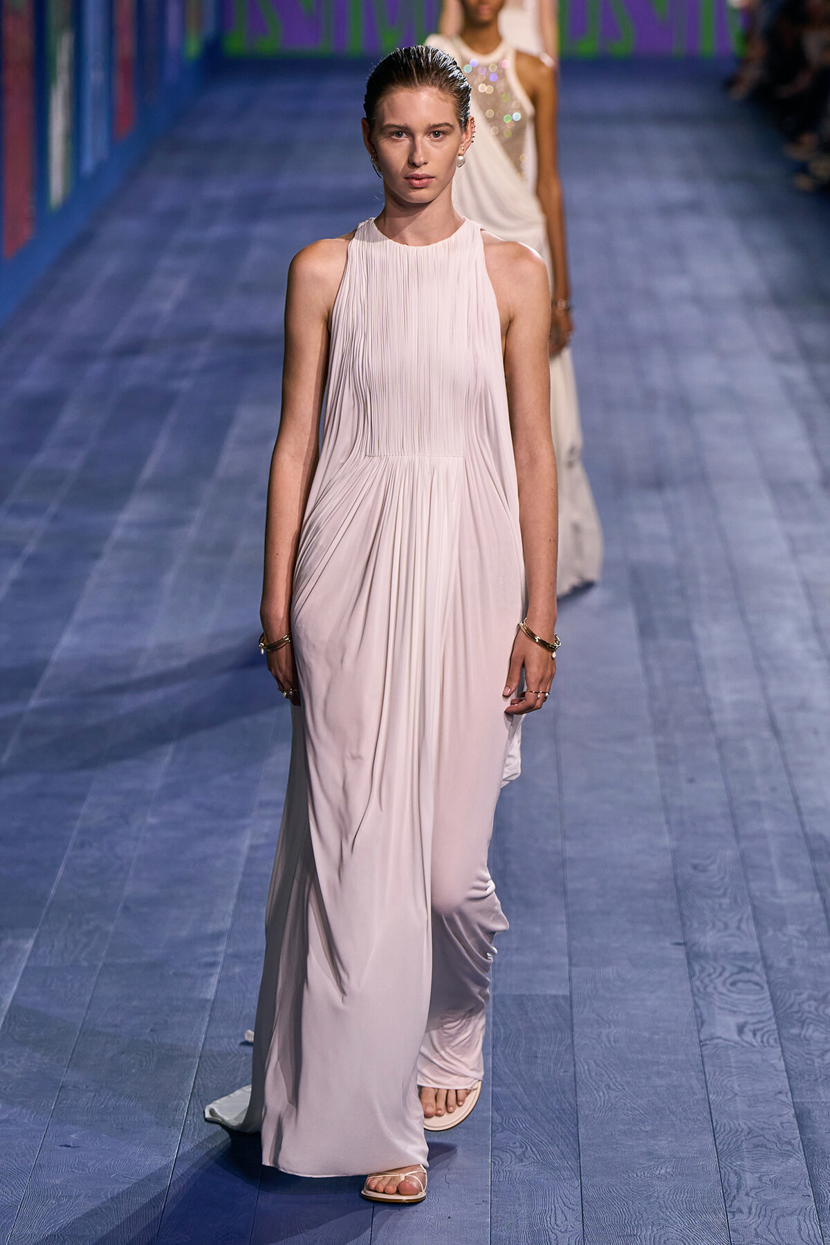 Model walking a pale pink, pleated halter-neck gown down a blue runway; minimal jewelry visible.