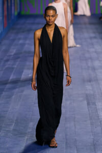 Model walking a runway in a sleeveless black draped halter gown. She wears braided hair and simple accessories.