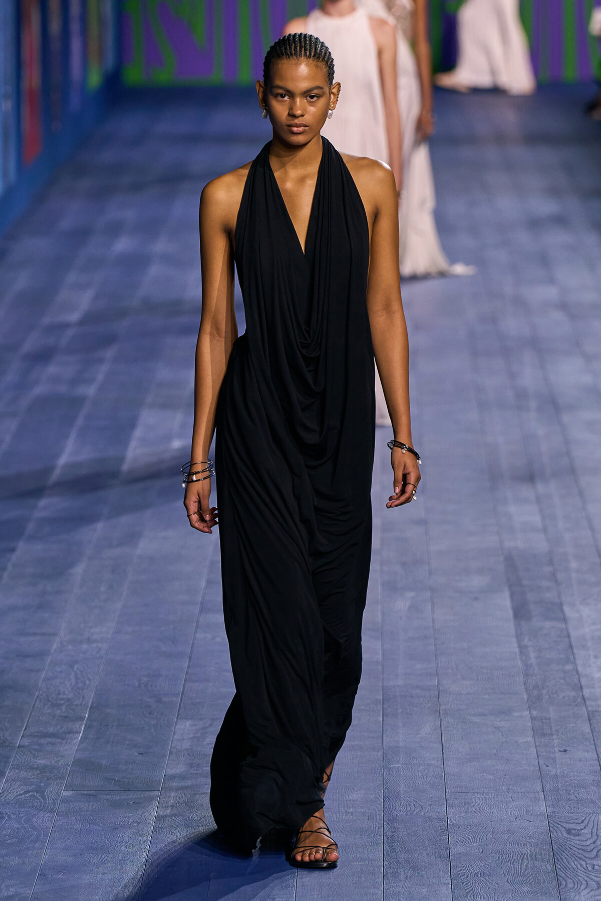 Model walking a runway in a sleeveless black draped halter gown. She wears braided hair and simple accessories.