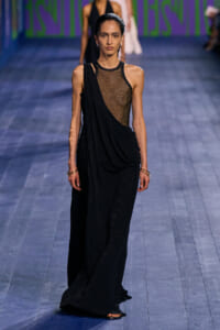 Model walking runway in a black draped gown with a metallic mesh bodice and one-shoulder drape detail.