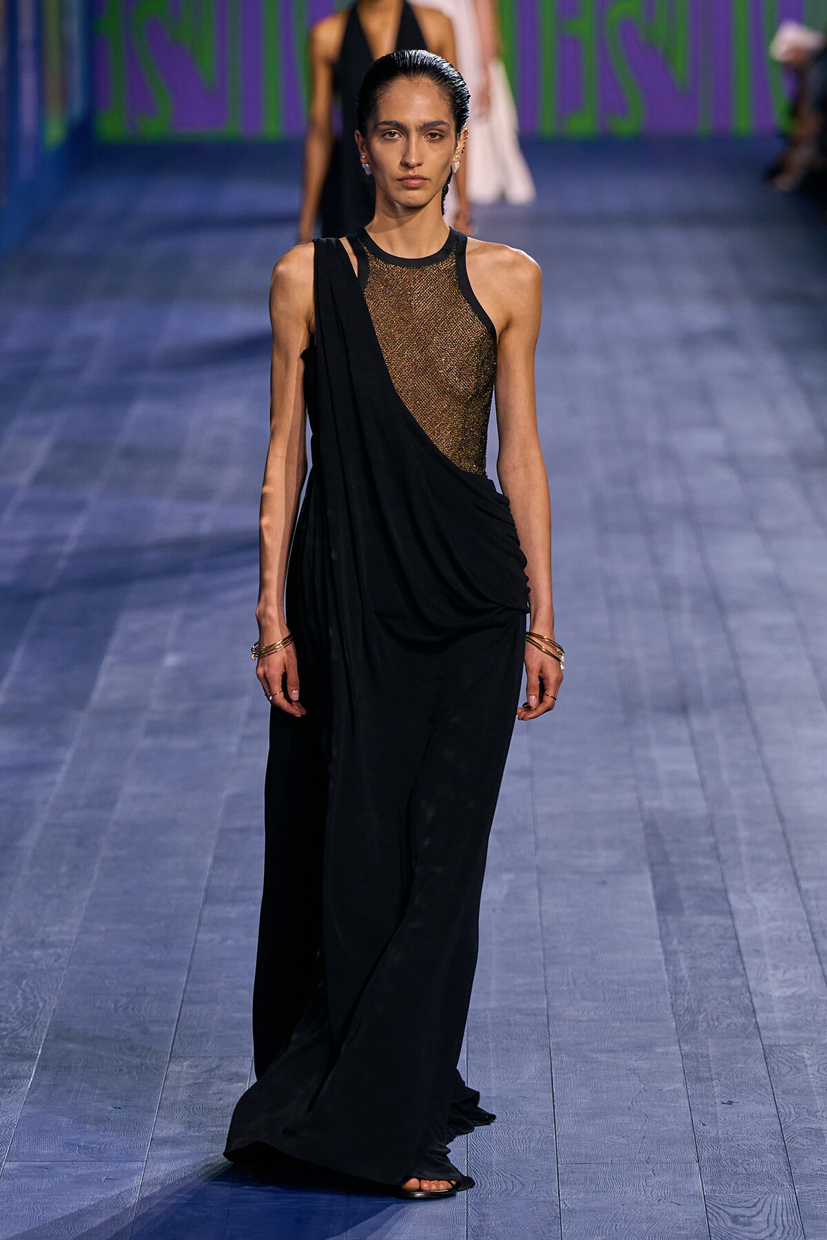 Model walking runway in a black draped gown with a metallic mesh bodice and one-shoulder drape detail.