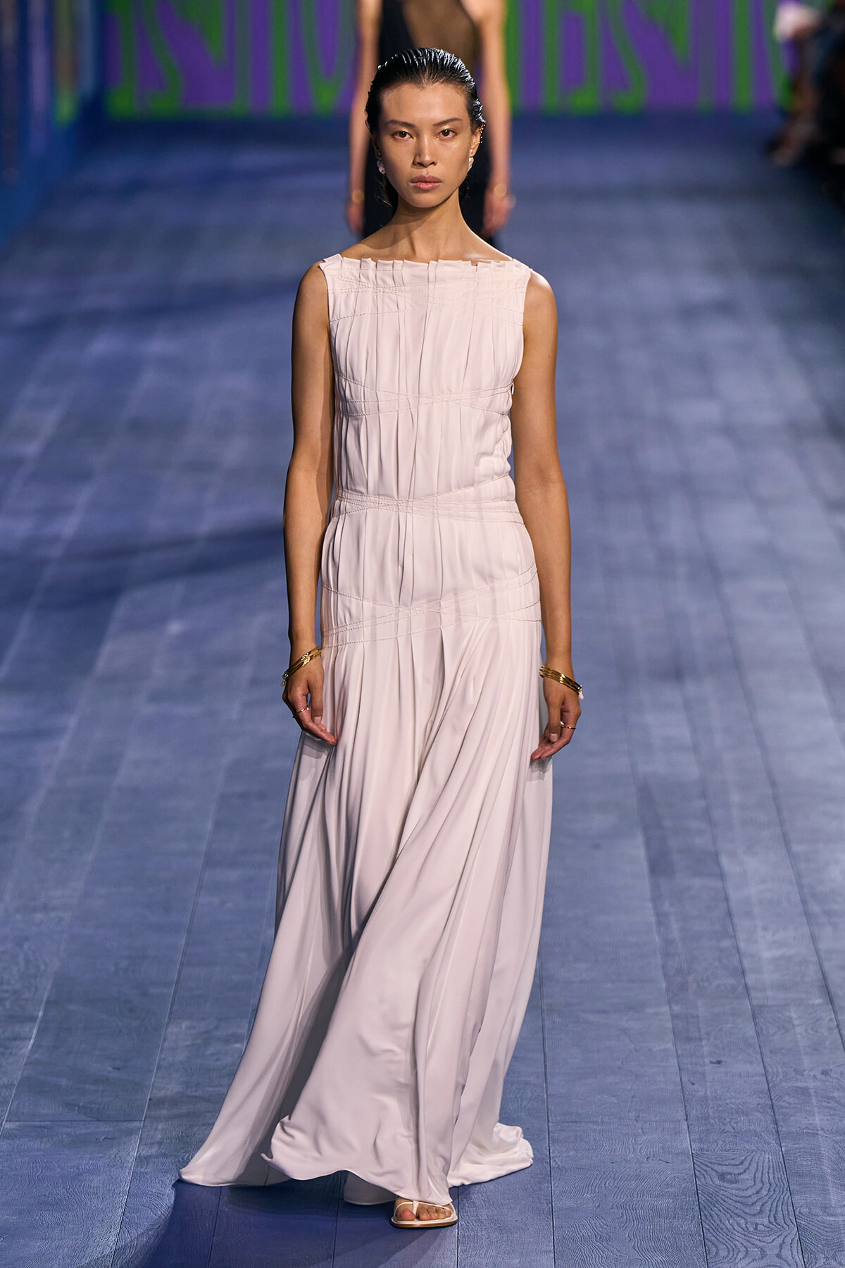 Model walking the blue runway in a pale pink sleeveless pleated floor-length gown.