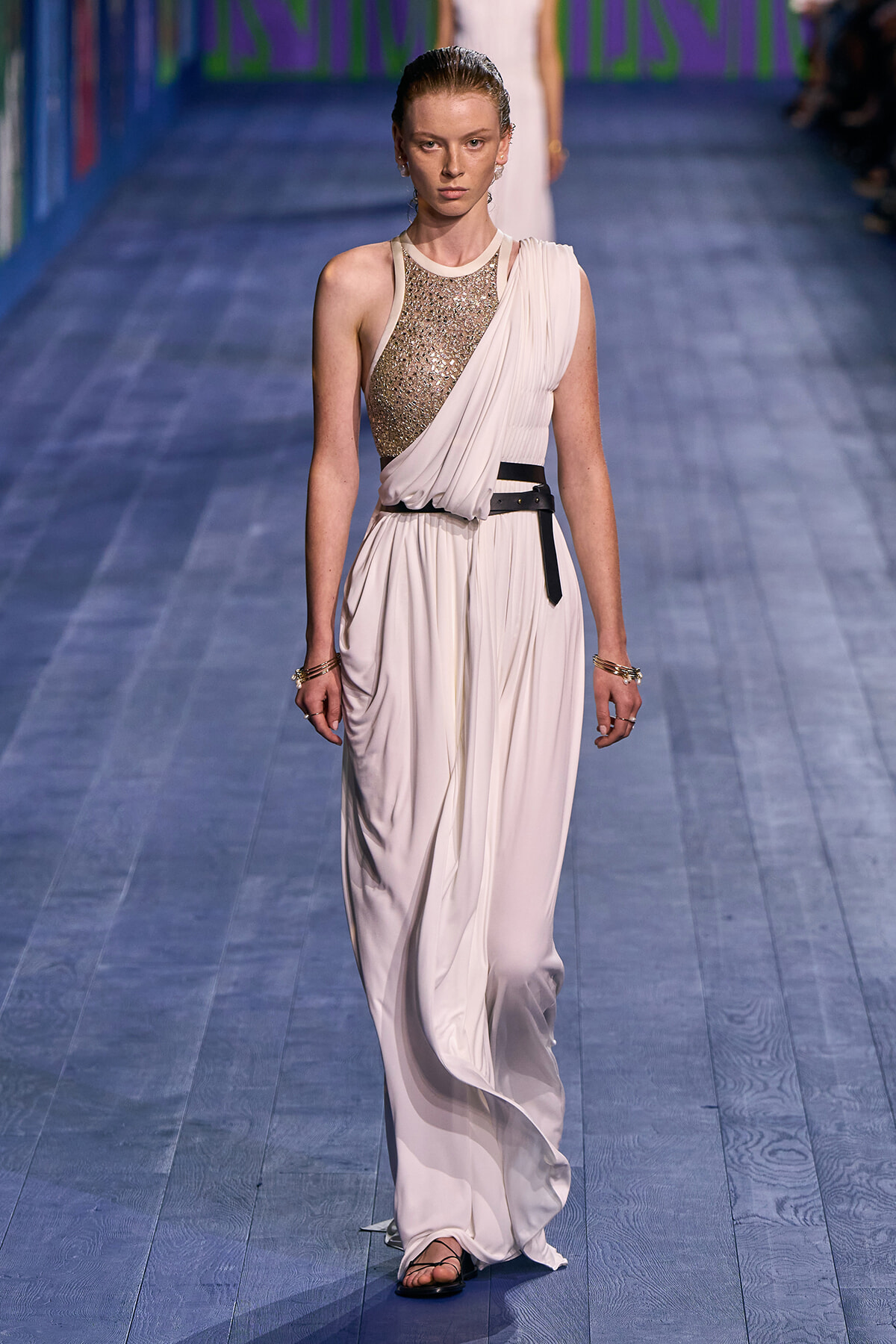 Model walking the runway in a cream, one-shoulder, draped gown with a gold sequined bodice and black belt.