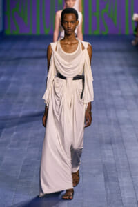 Female model walks the runway in a flowing ivory draped dress with a black belt and gladiator sandals, wearing minimal jewelry.