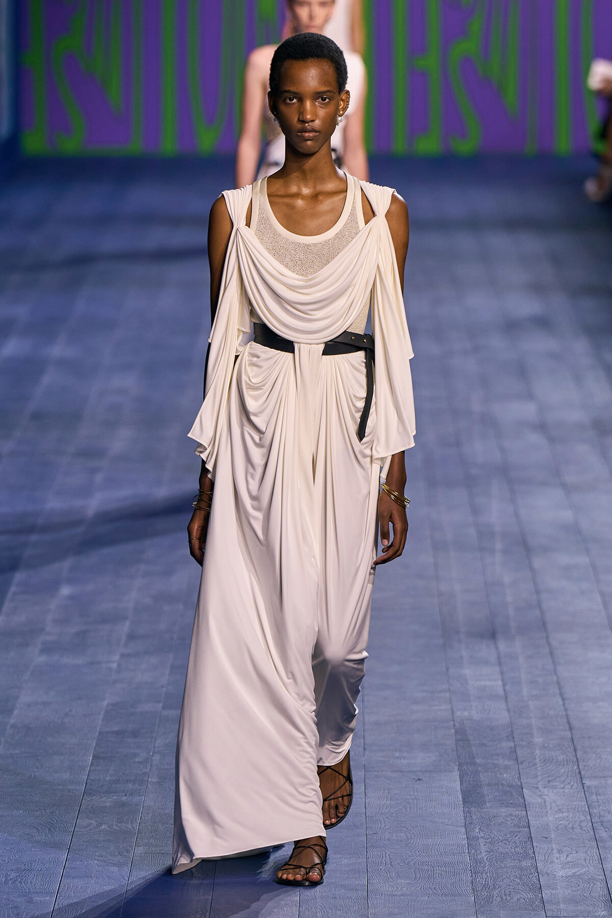 Female model walks the runway in a flowing ivory draped dress with a black belt and gladiator sandals, wearing minimal jewelry.