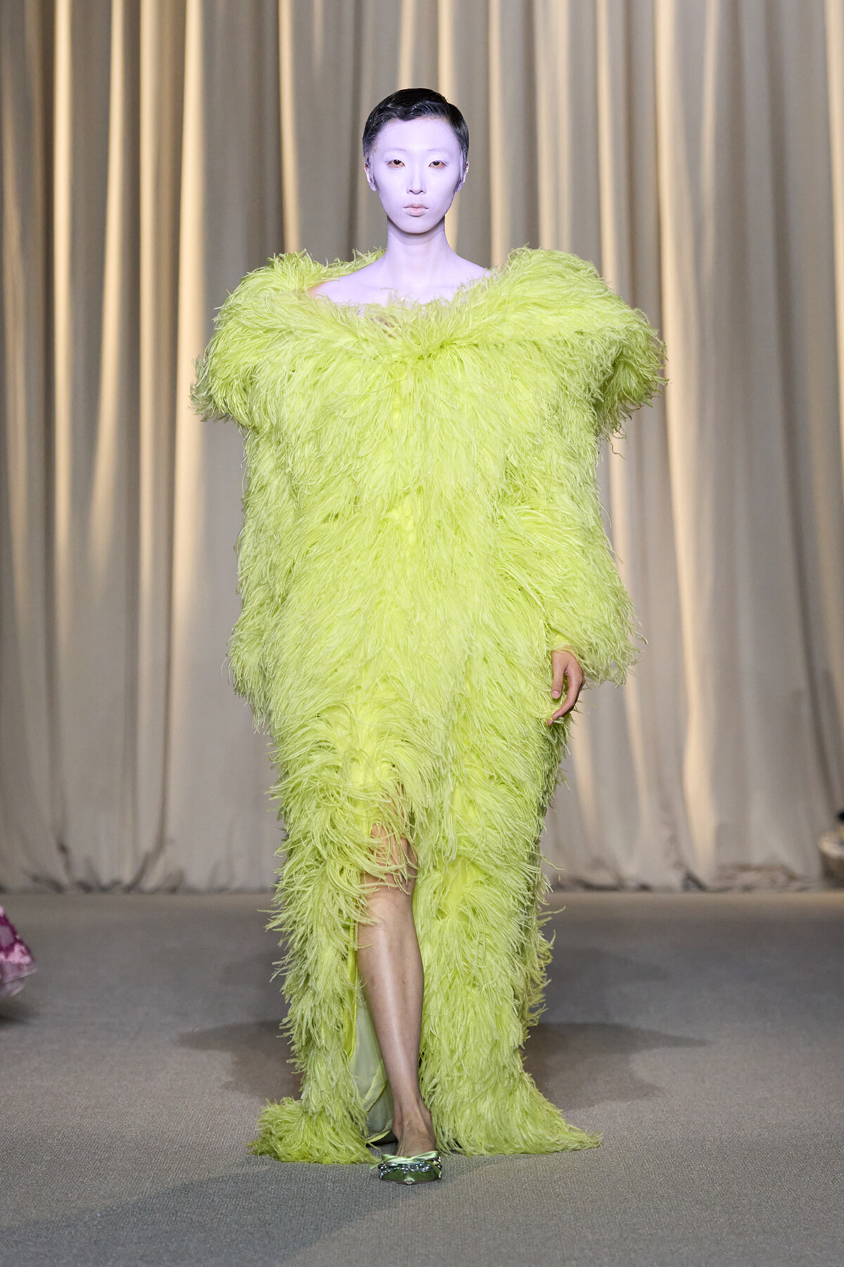 Model walking the runway in a neon lime feathered gown with oversized shoulders and a high slit, on a neutral backdrop.