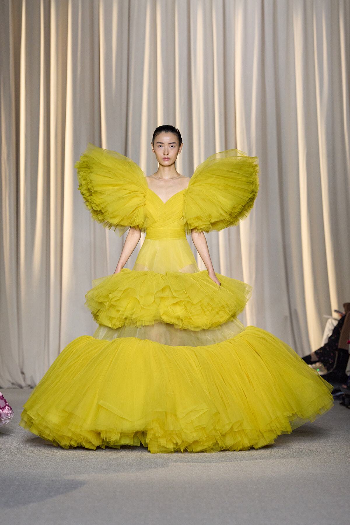 Model wearing a bright yellow voluminous gown with large ruffled shoulders and multi-tiered skirt on a runway.