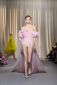 Model walks the runway in a pale pink off-the-shoulder bodysuit with oversized pink rose appliqués and a sheer cape, petals stuck to her skin and shoulders.