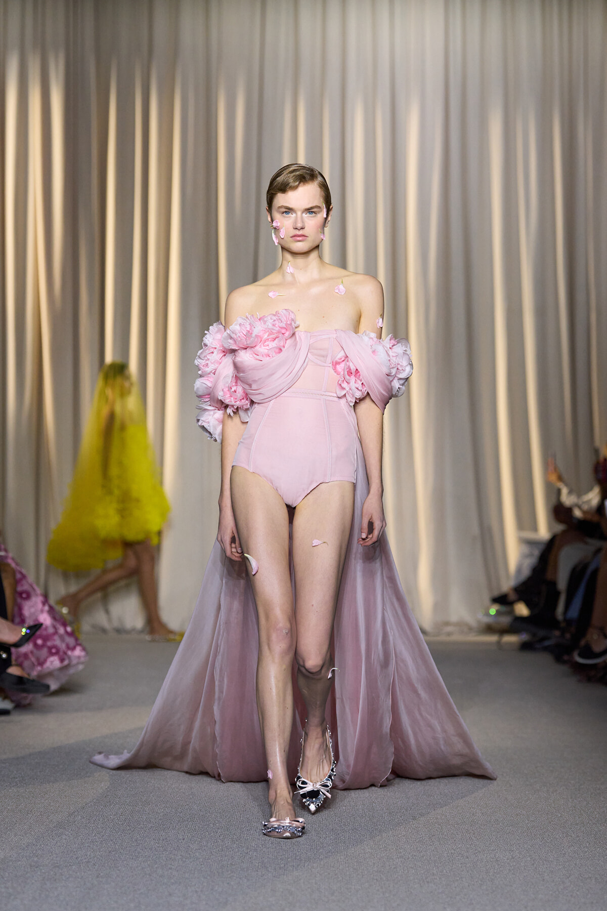 Model walks the runway in a pale pink off-the-shoulder bodysuit with oversized pink rose appliqués and a sheer cape, petals stuck to her skin and shoulders.