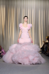Model walking a runway in a pale pink, voluminous tulle gown with ruffled shoulder accents and a tiered, fluffy skirt.