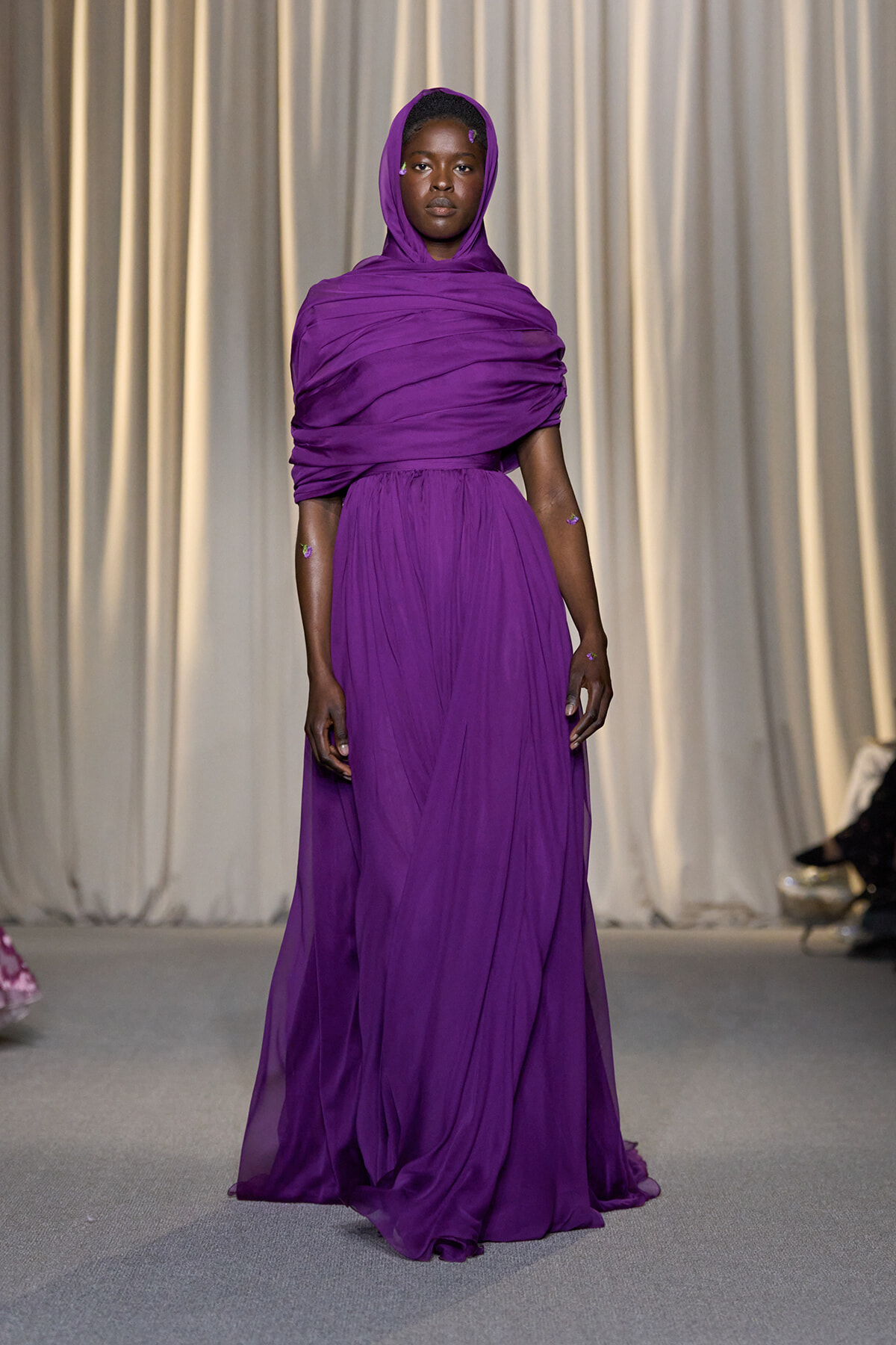 Model wearing a vivid purple draped gown with hood walking a runway on a fashion show