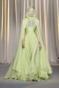 Model on a runway wearing a lime green gown with a sheer hooded cape and a bejeweled bodice.