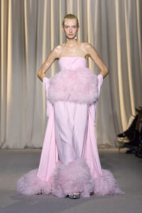 Model on a runway wearing a pale pink strapless gown with a large fluffy feather belt and feathered hem.