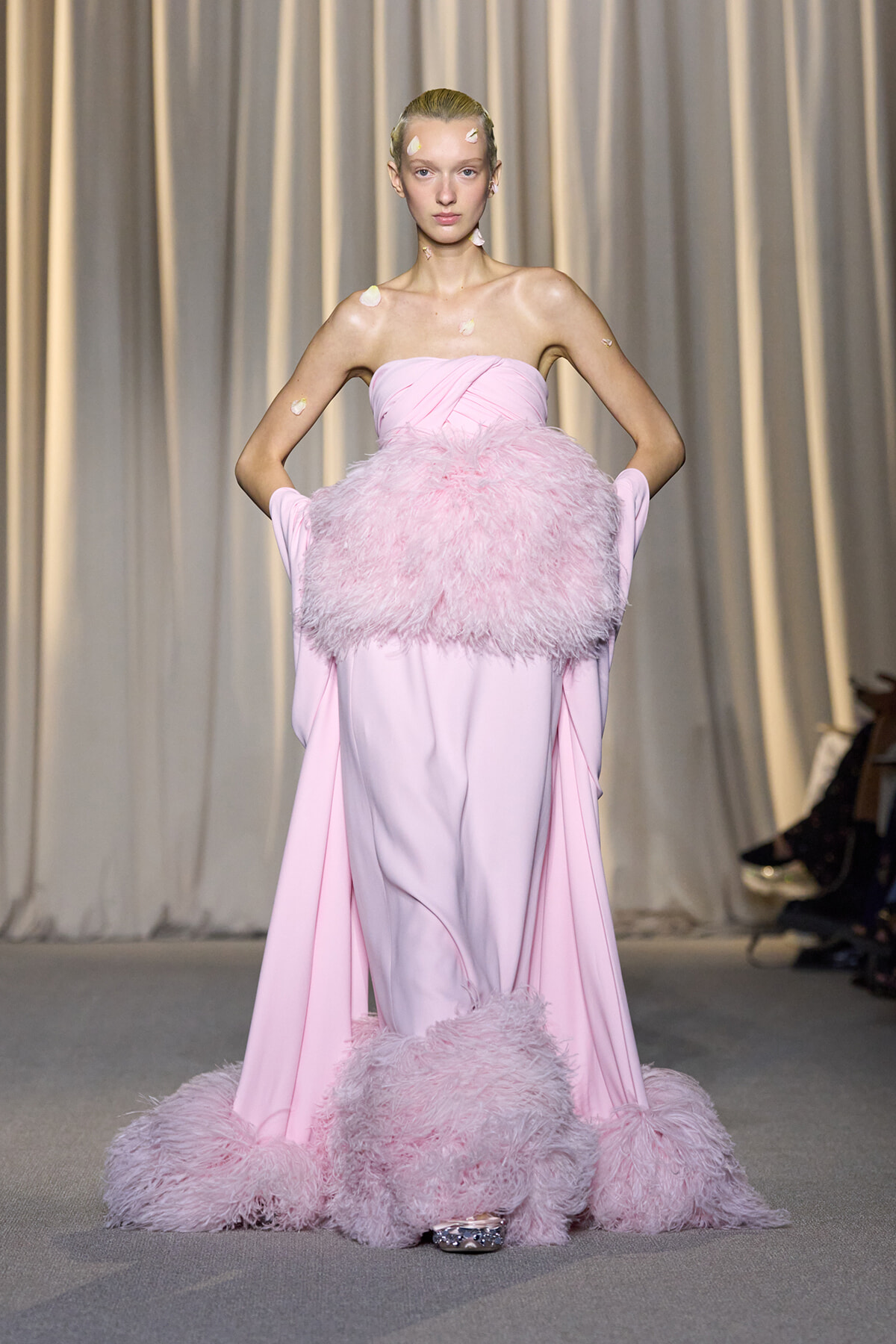 Model on a runway wearing a pale pink strapless gown with a large fluffy feather belt and feathered hem.