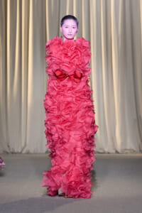 Model on a fashion runway wearing a floor-length pink gown with dense ruffles and a red bow at the waist, standing before a neutral curtain backdrop.
