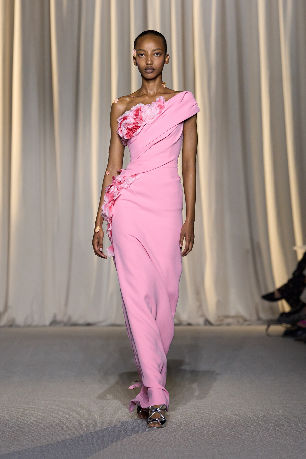 Model walking a runway in a pink one-shoulder gown with large floral appliqués along the bodice and hip.