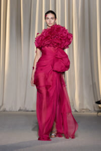 Model walking runway in a vivid fuchsia gown with oversized ruffled shoulder detail and draped skirt, neutral backdrop.