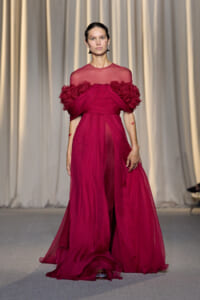 Model on a runway wearing a vivid magenta dress with a sheer bodice and large floral-embellished sleeves.
