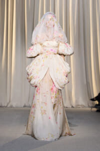 Model on a runway wearing a pale floral gown with oversized draped sleeves and a sheer floral veil.