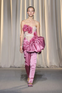 Model walking a runway in a pink satin gown with a large bow and floral bodice embellishments.