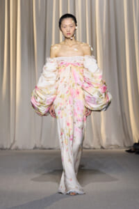Model on a fashion runway wearing an ivory off-the-shoulder gown with pink-yellow floral print and oversized puff sleeves.