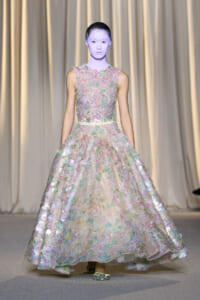 Model on a runway wearing a full-length pastel floral gown with iridescent petals and lavender-toned makeup, walking the catwalk.