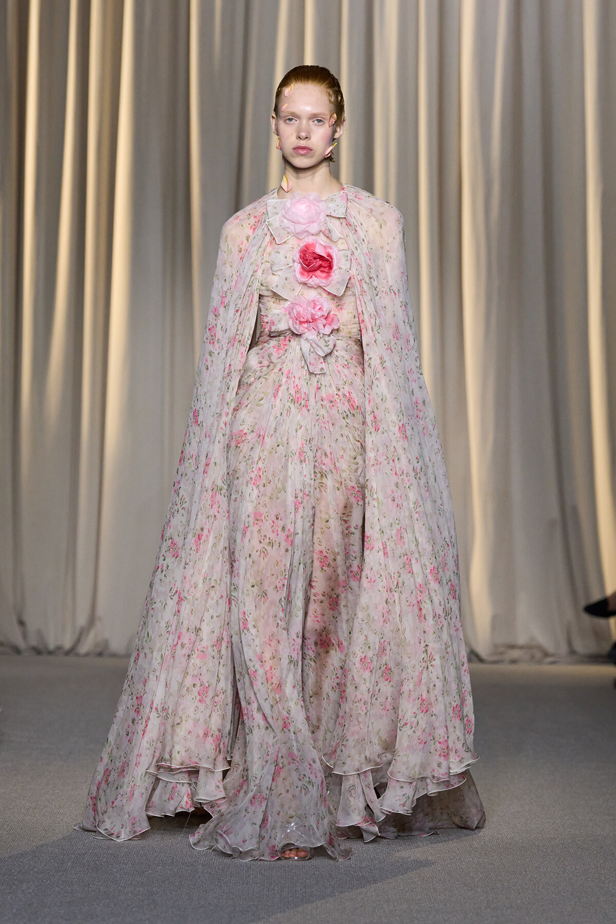 Model wearing a long, pale pink floral gown with a cape and large pink flower embellishments on the bodice, walking on a runway.