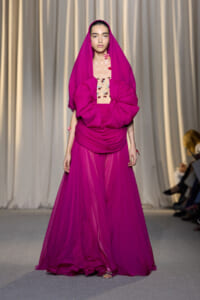 Model walks the runway in a magenta gown with a hooded draped cape and large bow at the waist, sheer skirt.