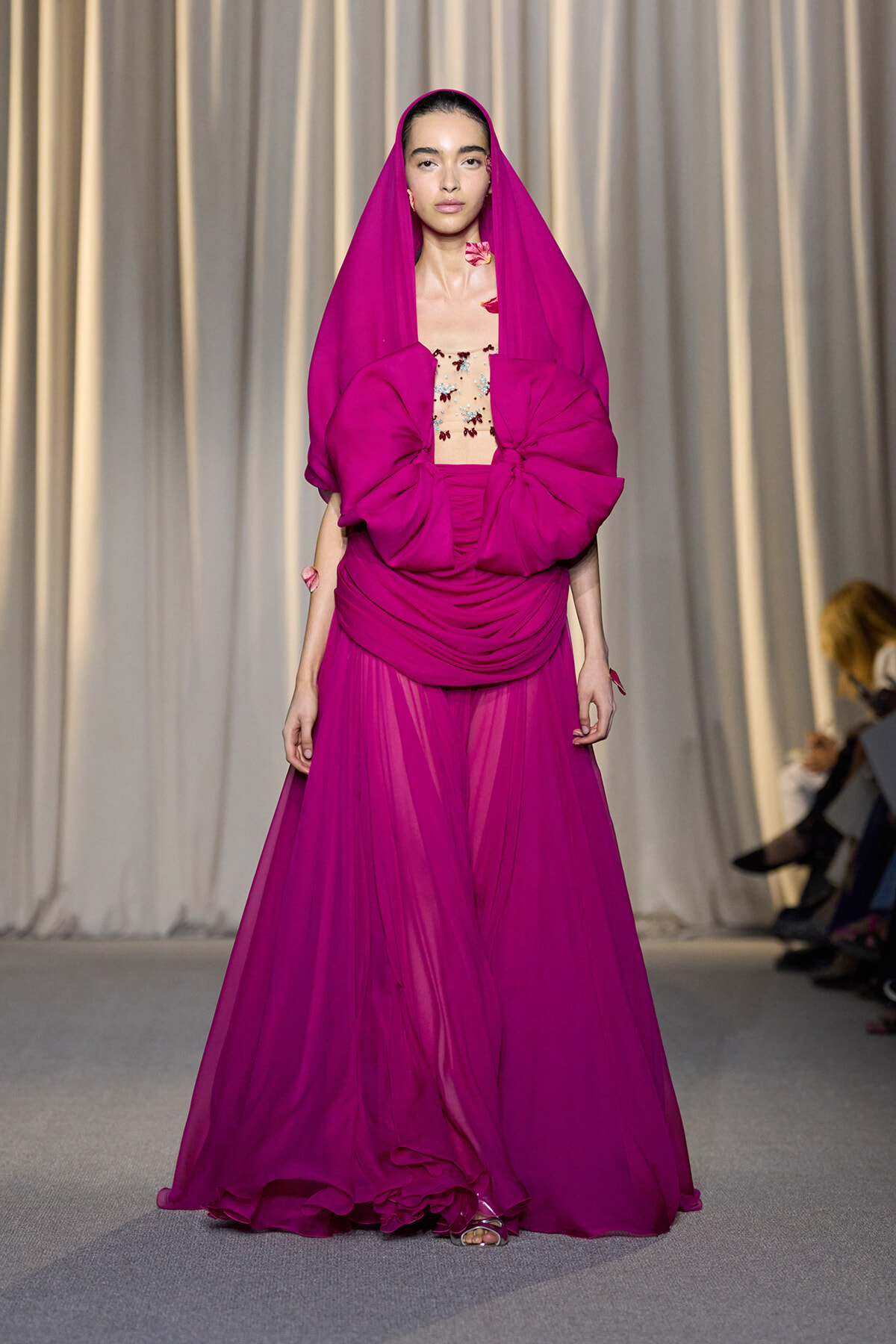 Model walks the runway in a magenta gown with a hooded draped cape and large bow at the waist, sheer skirt.
