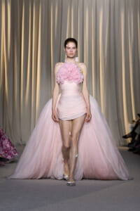 Model walking a pink, ruched mini dress with a large rose-hued flower bodice on a runway with a voluminous train behind her