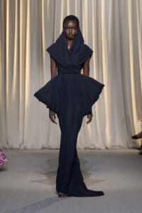 Model on a runway wearing a navy draped gown with a hooded overlay and a voluminous, wing-like skirt, walking toward the camera against a neutral backdrop.