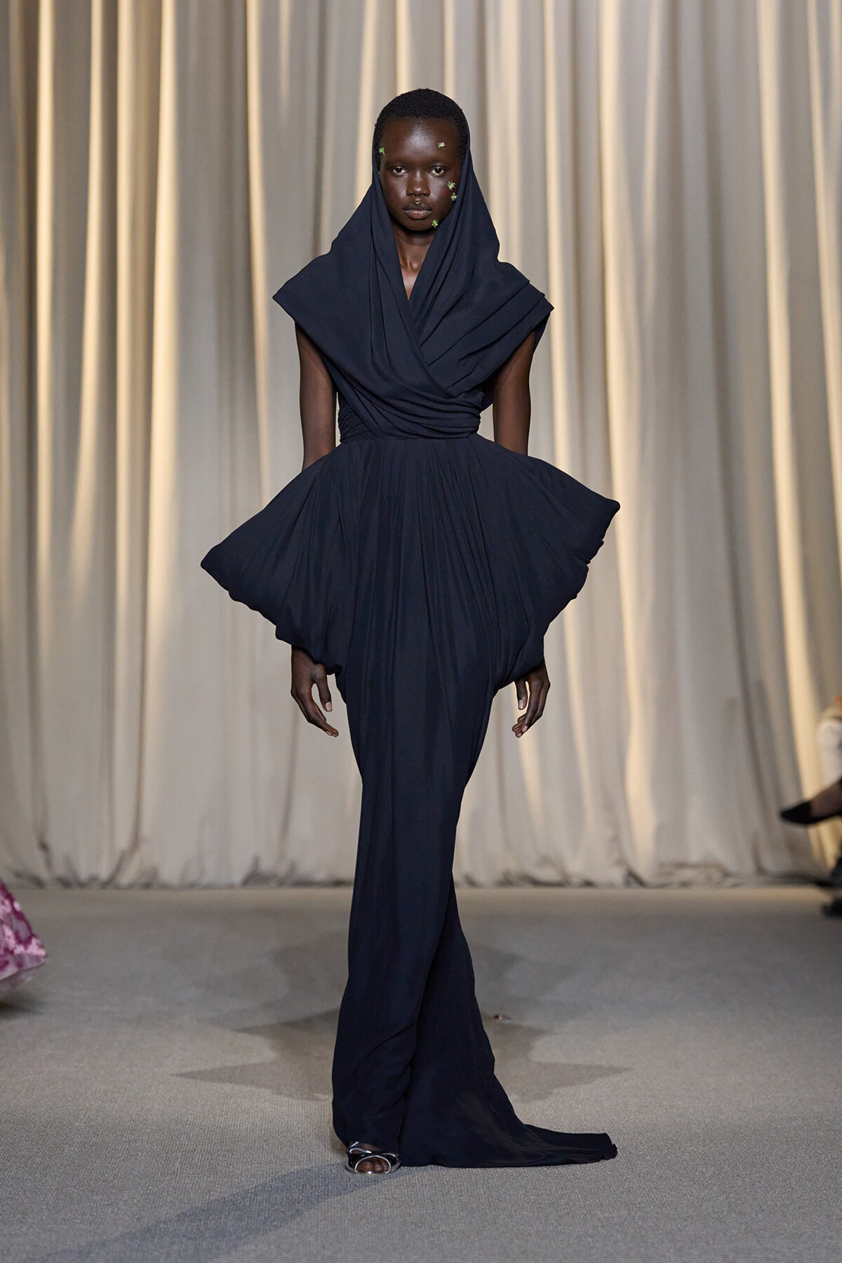 Model on a runway wearing a navy draped gown with a hooded overlay and a voluminous, wing-like skirt, walking toward the camera against a neutral backdrop.