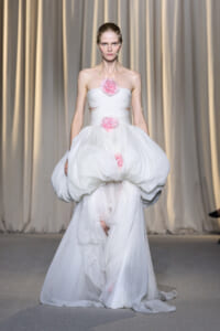 Model walking a runway in a strapless white gown with a voluminous, draped skirt and pink flower embellishments at the bodice and waist.