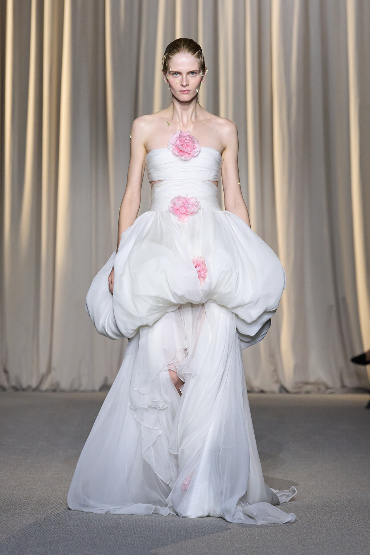Model walking a runway in a strapless white gown with a voluminous, draped skirt and pink flower embellishments at the bodice and waist.