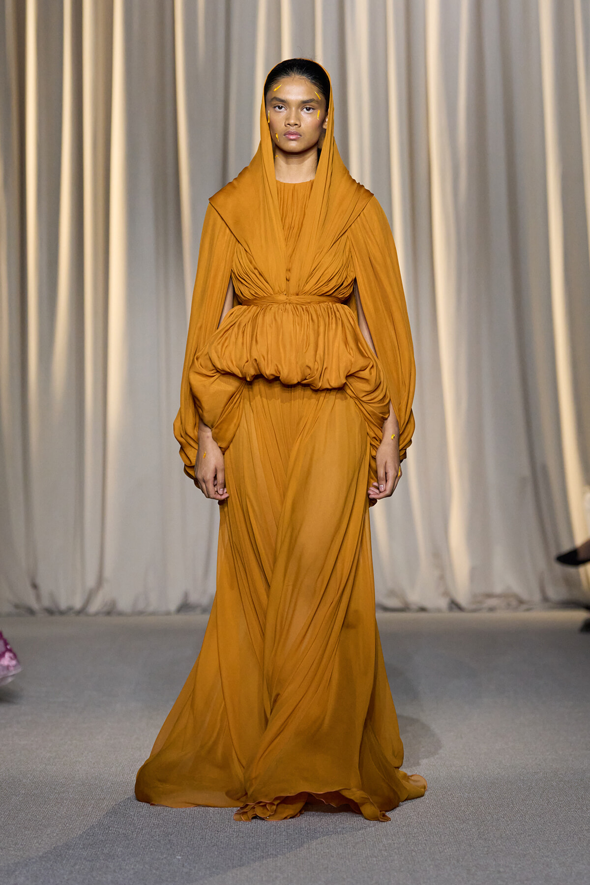 Model walking a runway in a mustard-orange, draped gown with a hooded cape and a cinched, voluminous waist.