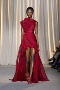 Model walking a runway in a burgundy red one-shoulder gown with a large floral shoulder embellishment and a sheer, ruffled high-low train.