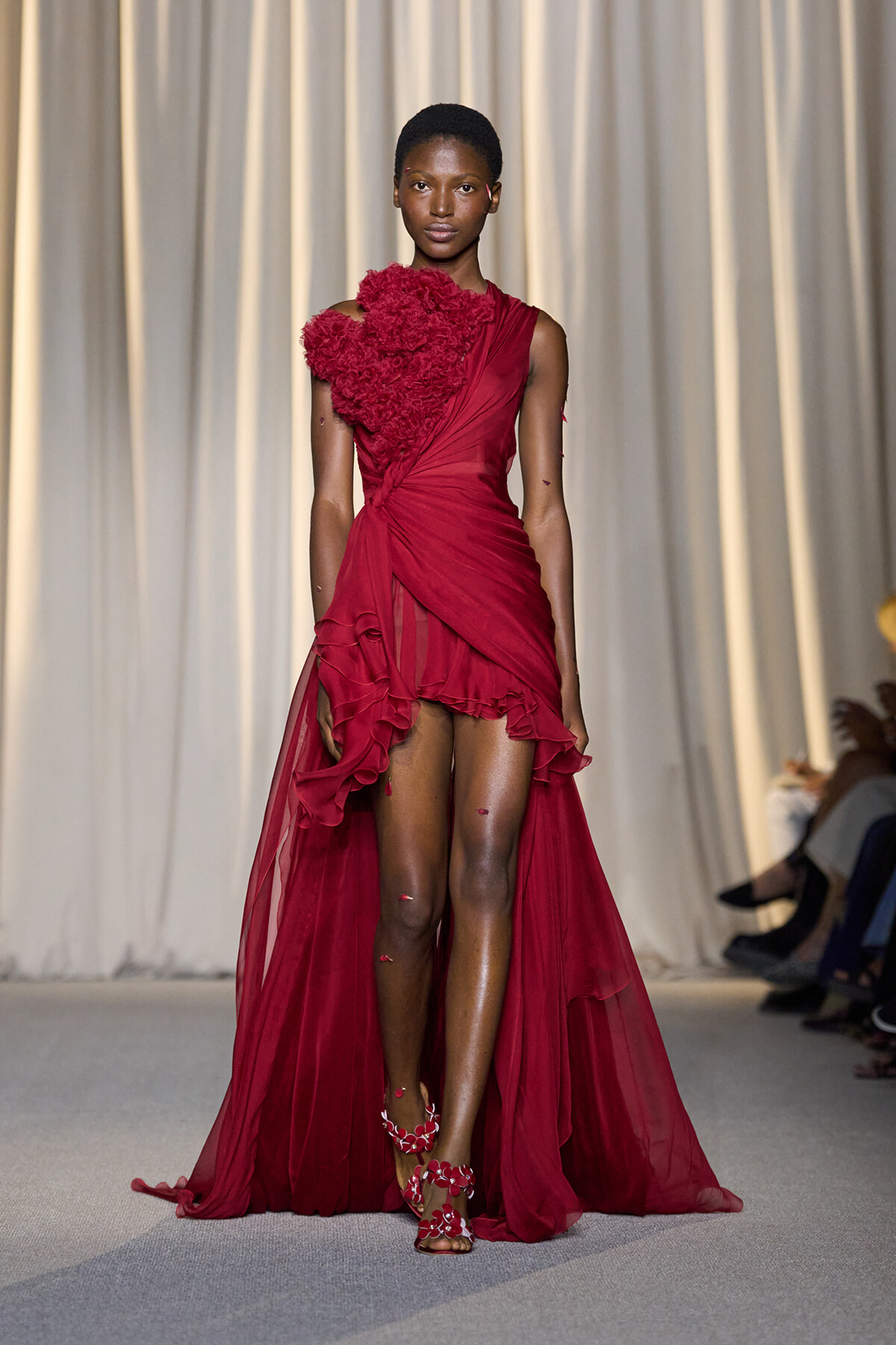 Model walking a runway in a burgundy red one-shoulder gown with a large floral shoulder embellishment and a sheer, ruffled high-low train.