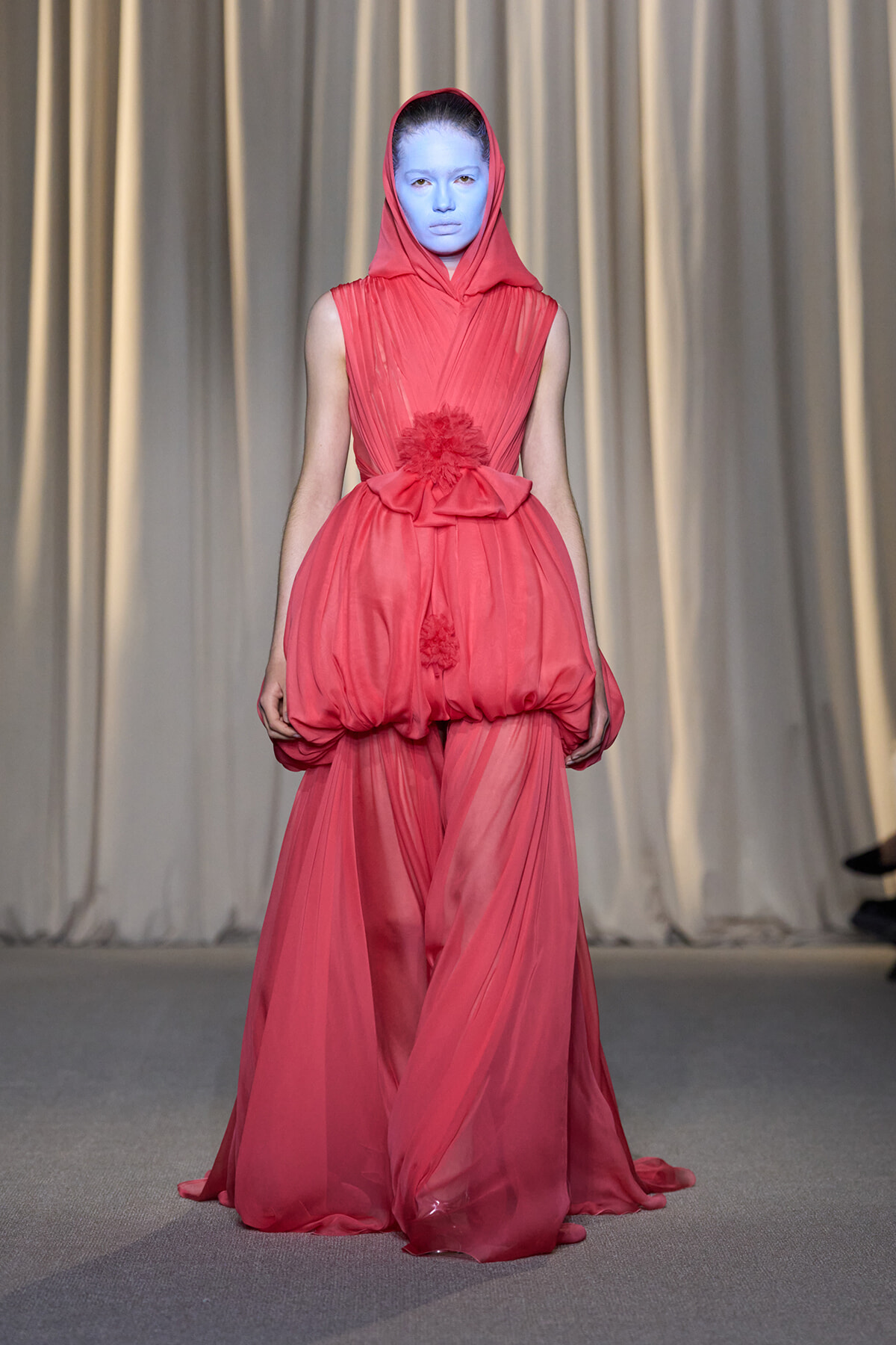 Runway model in a coral-pink hooded gown with a voluminous bubble skirt and blue face makeup.