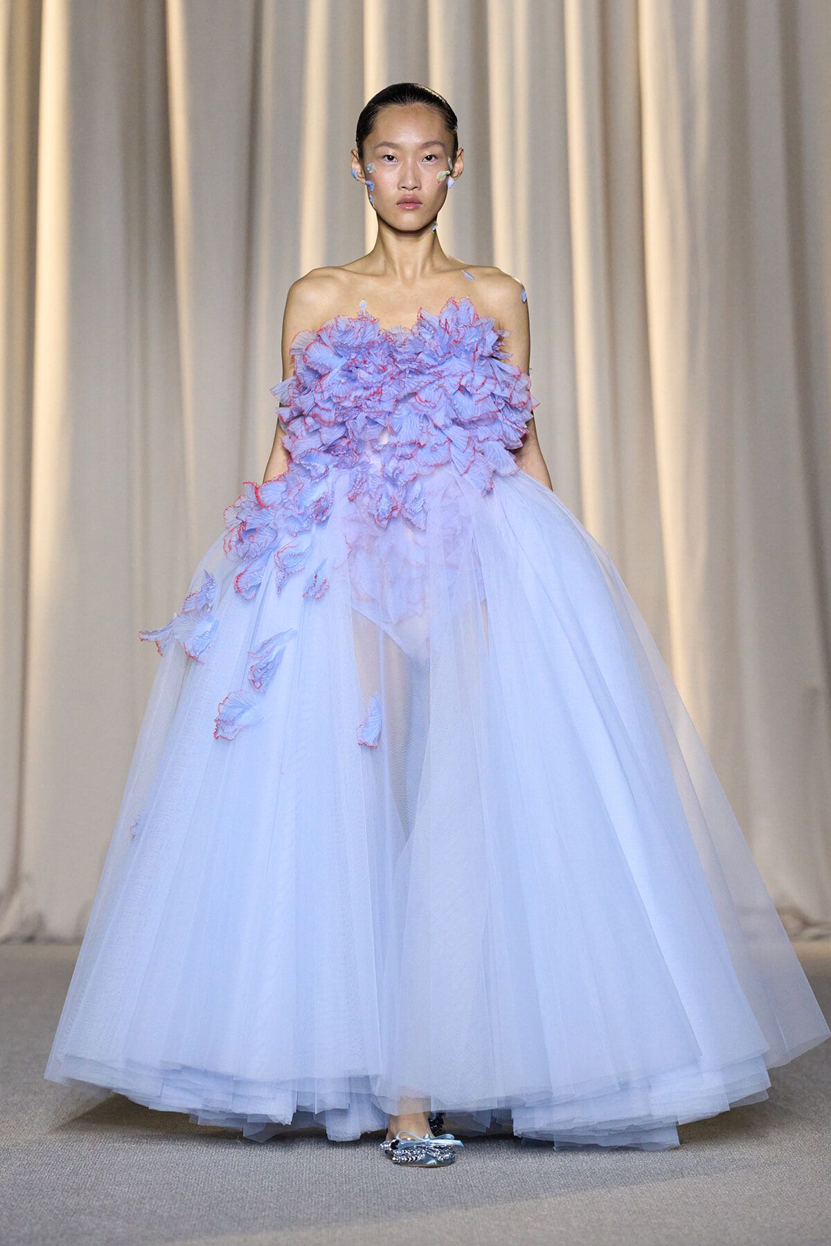 Model on a runway wearing a pale blue off-shoulder tulle ball gown with blue-pink floral appliqués cascading from the bodice.