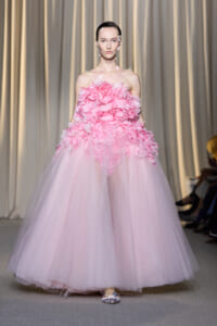 Model walking the runway in a pink off-shoulder ball gown with layered flower appliqués and a full tulle skirt on a fashion show runway.