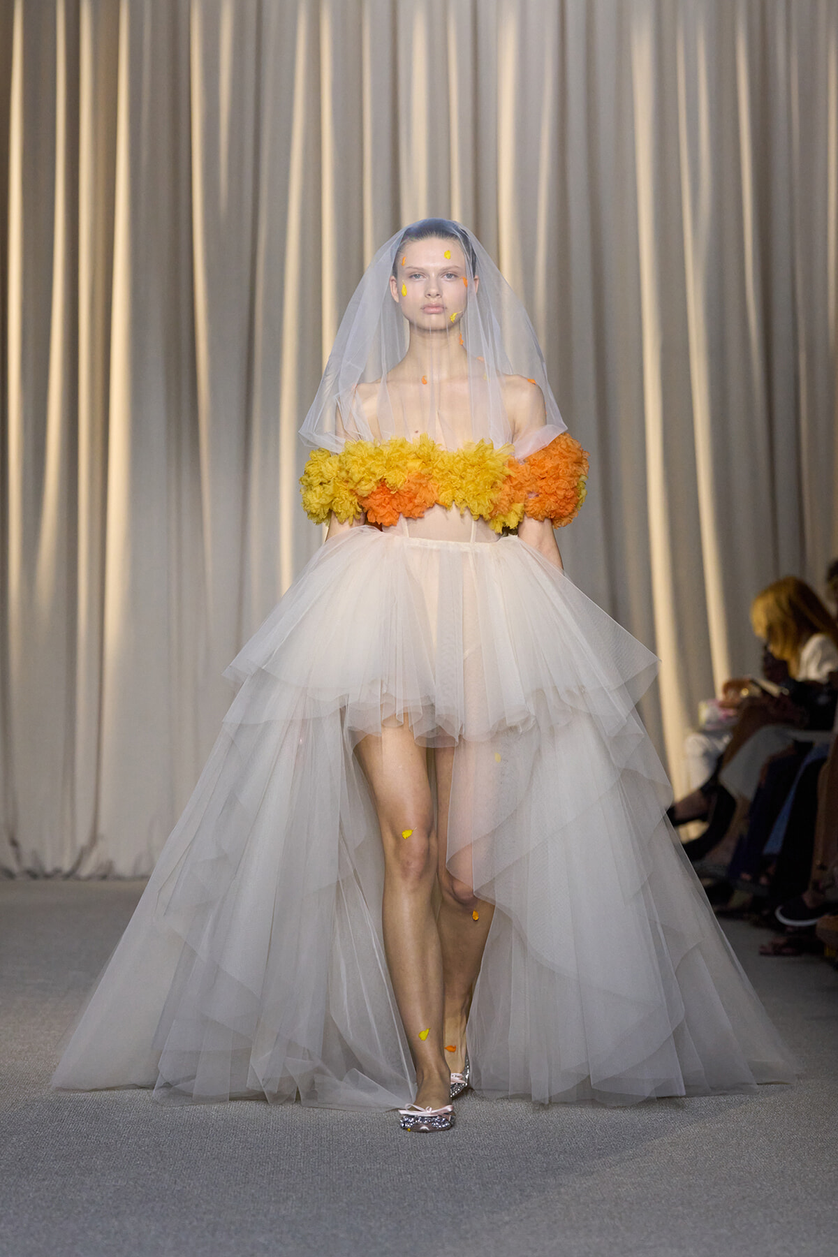 Model walks the runway in a white voluminous tulle gown with an off-shoulder yellow–orange floral bodice and a sheer veil.
