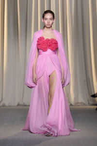 Model in a pink strapless gown with a voluminous floral ruffle bodice and sheer cape walking the runway.