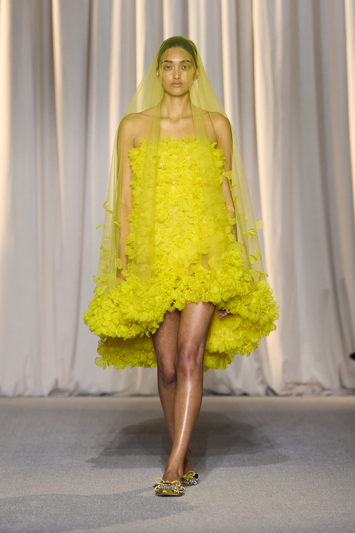 Model walking the runway in a bright yellow floral strapless mini dress with a matching yellow veil. She wears embellished yellow flats and stands against a neutral backdrop.