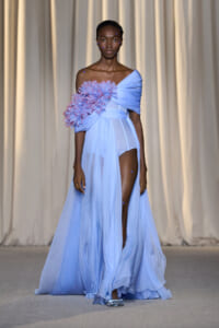 Model walking a fashion runway in a light blue off-shoulder gown with a large pink-blue floral cluster on the bodice and sheer skirt with a high slit.