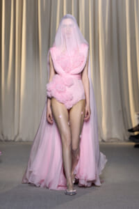 Model walks the runway in a pink, structured bodice gown with a sheer veil and long tulle cape.