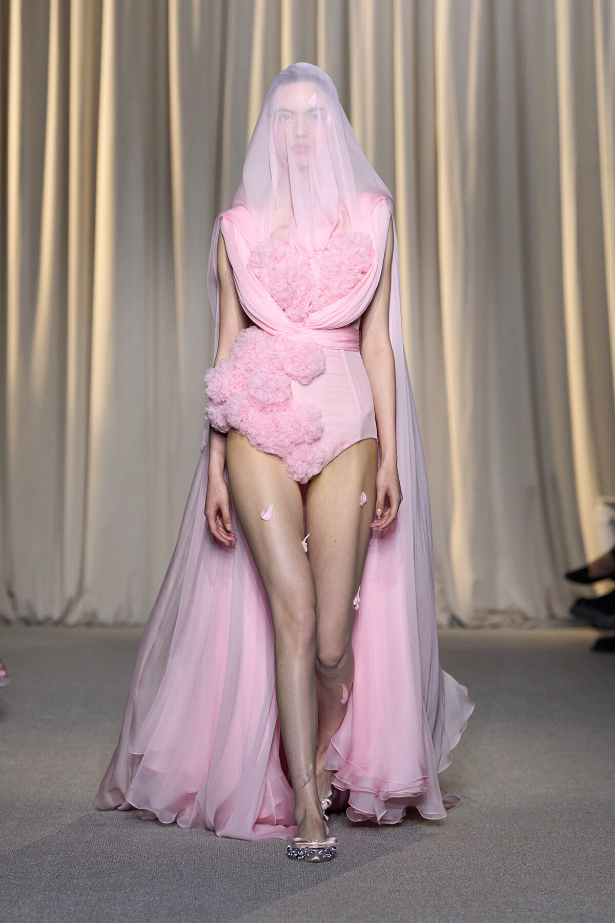 Model walks the runway in a pink, structured bodice gown with a sheer veil and long tulle cape.